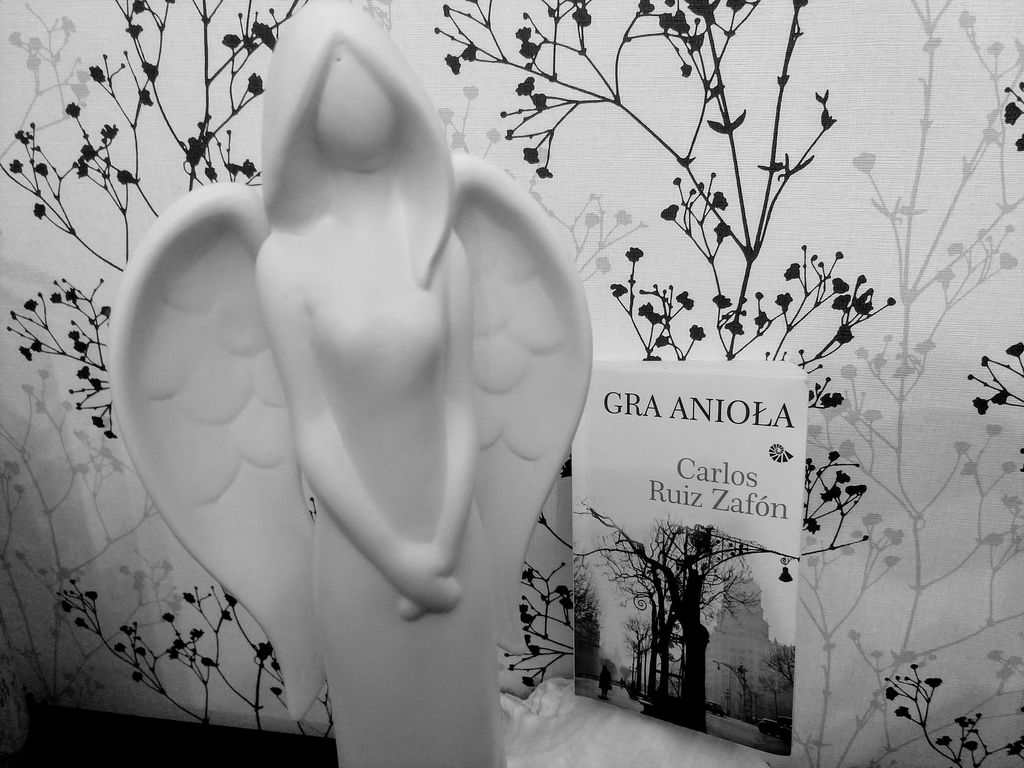 Biało-czarne zdjęcie przedstawiające okładkę książki "Gra anioła" Zafona obok białej figurki anioła
